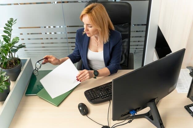 An employee reviewing a benefits document at their desk, highlighting key points about tuition reimbursement. Their laptop displays the company's HR portal. Natural light fills the workspace.
