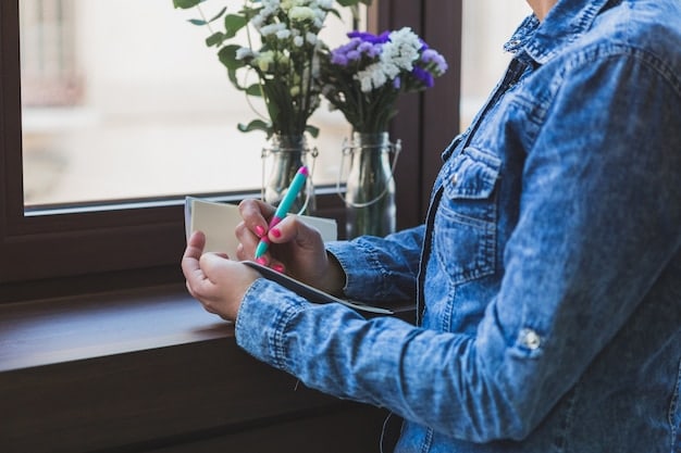 A close-up shot of a person writing in a gratitude journal by a window with natural light. The journal is open, and the person is using a pen to write, with a cup of tea and a small plant nearby, creating a cozy and mindful atmosphere.