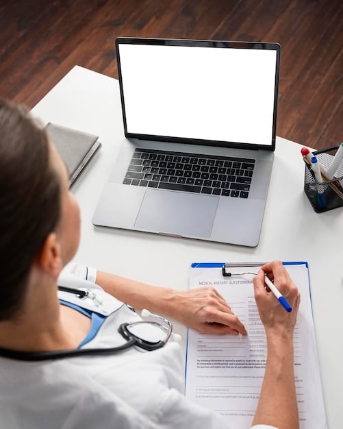 A person sitting at a desk reviewing documents related to Medicaid eligibility, with a laptop displaying a state Medicaid website. The focus is on the documents and the laptop screen.